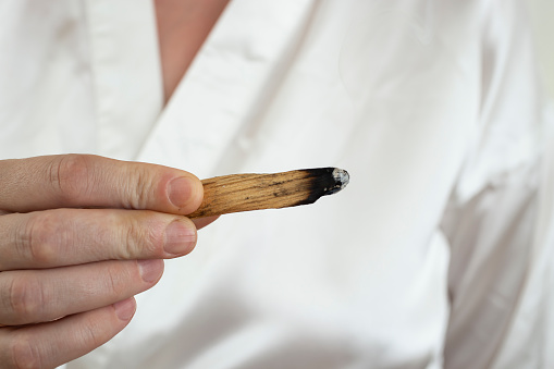 woman hand holding a stick of sacred palo santo wood, burning. white satin background.