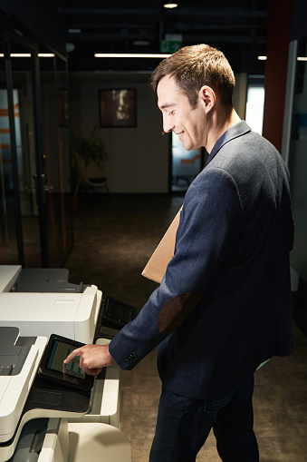 Desk worker standing near printer and clicking on its display while adjusting settings