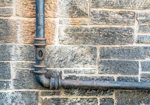 An old waste pipe on the exterior of a stone built old-fashioned building, showing signs of wear and corrosion.