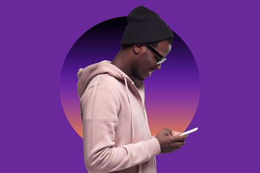 Side view of smiling millennial black guy in pink hoodie, hat and shades using mobile phone, checking social media, sending message against purple background with colorful gradient circle
