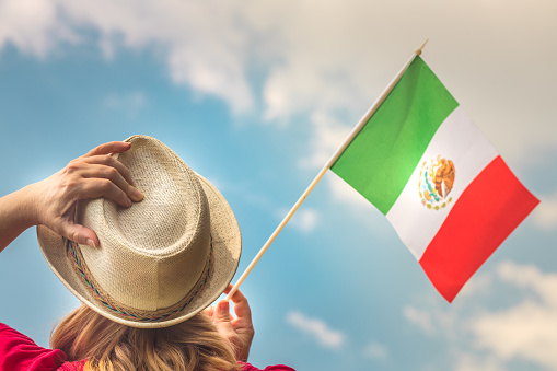 A young woman proudly raises the flag of Mexico above her head against the sky, The other hand holds up her hat, Concept for national holidays and patriotic events