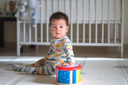 Mixed race baby boy playing with a toy drum in the bedroom while sitting on a baby safe soft playmat on the floor at home. 10 months old infant playing at home