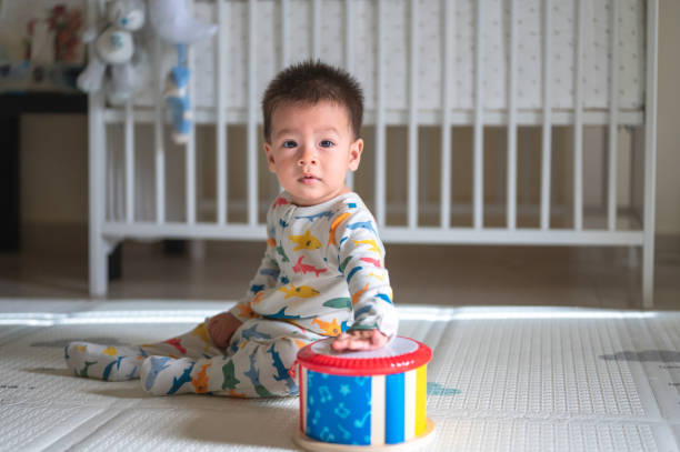 Baby boy playing with a toy drum in the bedroom while sitting on a baby safe soft playmat on the floor at home Mixed race baby boy playing with a toy drum in the bedroom while sitting on a baby safe soft playmat on the floor at home. 10 months old infant playing at home toddler hitting stock pictures, royalty-free photos & images