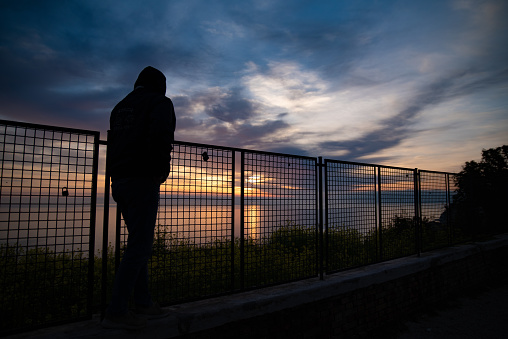 Silhouette of a Lonely Man Walking on Coastal Road in Sunrise over Sea Lights