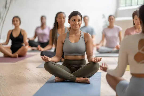 A large group of diverse adults participate together in a meditation class.  They are each seated on a yoga mat with their legs crossed and their hands on their knees as they focus on their breathing.