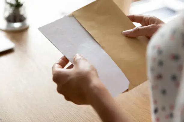 Hands of woman receiving letter, invitation, notification, postcard, taking out document for reading, opening envelope with blank folded paper at work desk. Mail concept. Close up, cropped shot