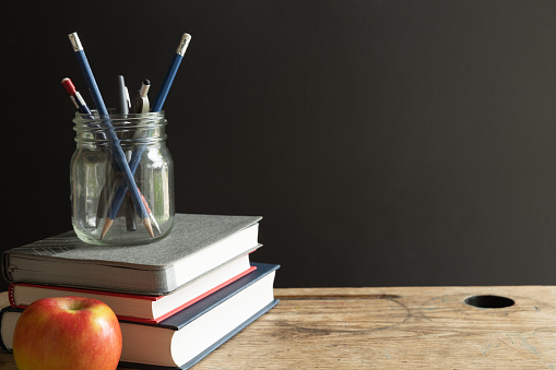 Stack of red, navy blue and grey books with a glass jar filled with pens and pencils on top of the books with a red apple on a vintage wood school desk with copy space in black background