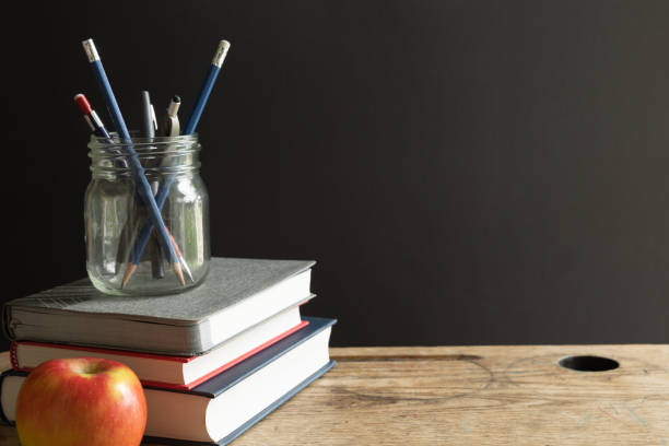 Books, jar of pens and red apple on vintage desk with black background stock photo