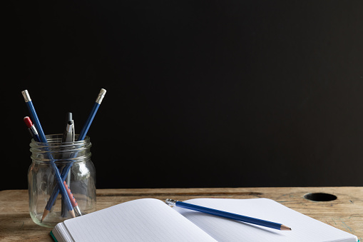 Open blank notebook with white lined pages laying on a vintage wood school desk with a jar of pencils with a black background and copy space