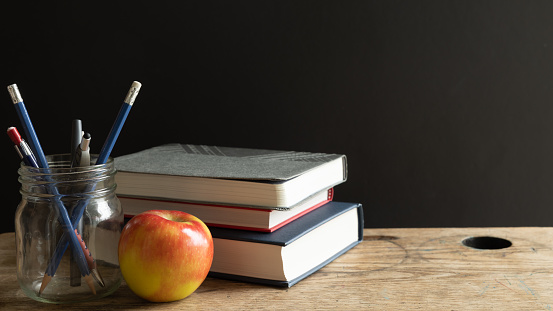 Glass jar filled with pens and pencils, red apple and a stack of navy blue, red and grey books on a vintage wood school desk with copy space in black background