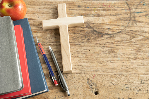 Wood Christian cross laying on a vintage wood desk with a stack of grey, red and navy blue books, a red apple and a pen and pencil, shot from above with copy space