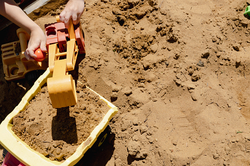 Hands of child playing with digger and yellow dump truck in sand box with copy space shot from above