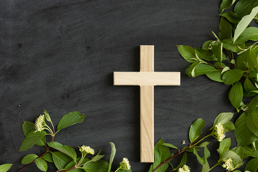 Small wood christian cross on a black chalkboard background with a border of branches with green leaves with copy space shot from above