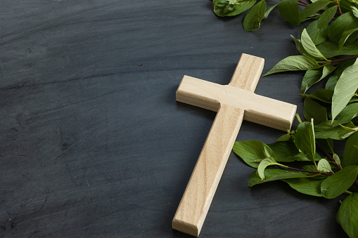 Small wood christian cross on a black chalkboard background with a border of branches with green leaves with copy space