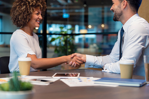 Business man and woman shaking hands in the office. There is a paperwork and technology on the table with some coffee. Both are happy and smiling