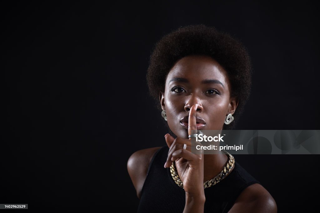 Closeup of beautiful young dark-skinned woman with finger on her lips showing shhh silence gesture on black background. Closeup of beautiful young african american dark-skinned woman with finger on her lips showing shhh silence gesture on black background. 20-24 Years Stock Photo