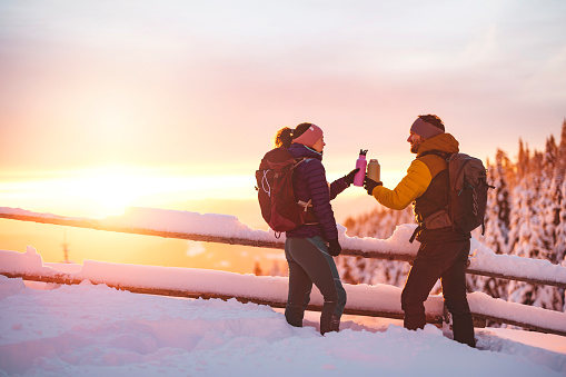 Mountaineering coupe giving cheers with their water bottles while on a winter hike in snowy mountains. Sun rising in the background making the sky pink.