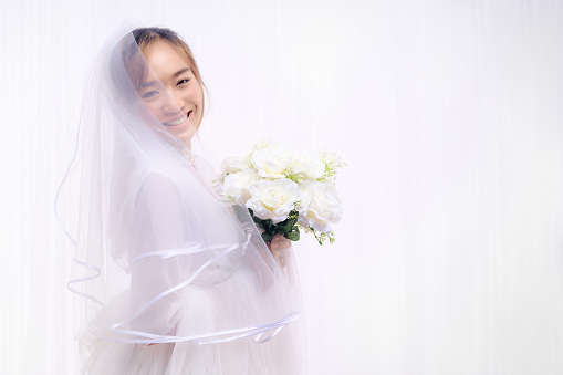 studio portrait of asian bride in white gown with veil holding flower bouquet on white background