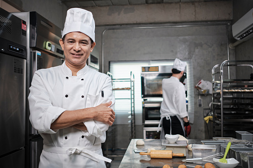 Professional senior Asian male chef in white cooking uniform, food occupation looks at camera, arms crossed, cheerful smile, expertise in commercial pastry culinary jobs, catering restaurant kitchen.