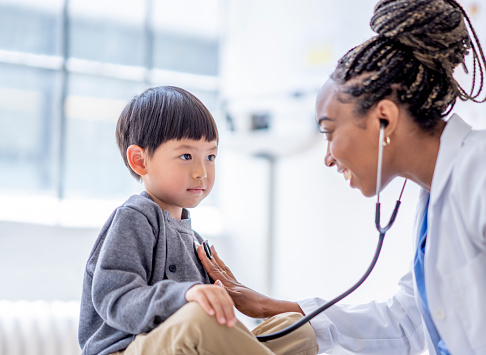 A female doctor leans in with her stethoscope as she listens to a little boys heart.  She is wearing a white lab coat and is smiling at the young boy who is seated cross legged on the exam table.