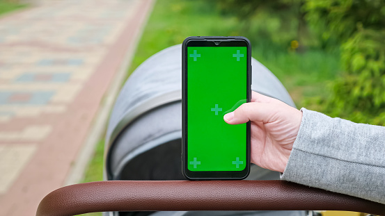 Woman touches green chromakey screen of phone put on stroller handle on blurred background. Mother holds electronic device on walk with baby in park closeup