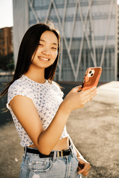 istock Young Asian woman looking at camera and smile while holding a mobile phone outdoors on the street. 1402995600