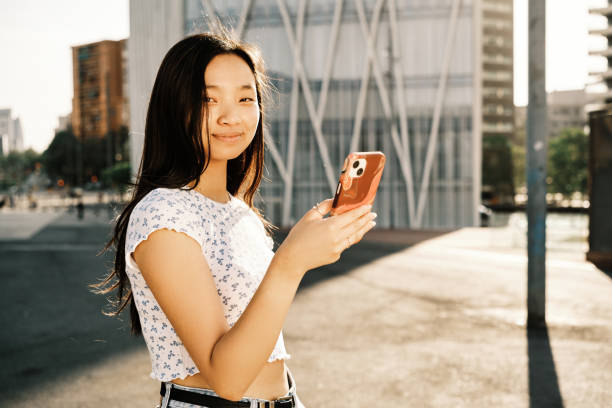 istock Young Asian woman looking at camera while using her mobile phone outdoors on the street. 1402995601