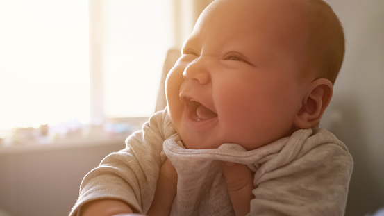 Cute newborn girl with plump cheeks smiles widely feeling happily at home. Mother holds baby with hands against bright window in bedroom close view