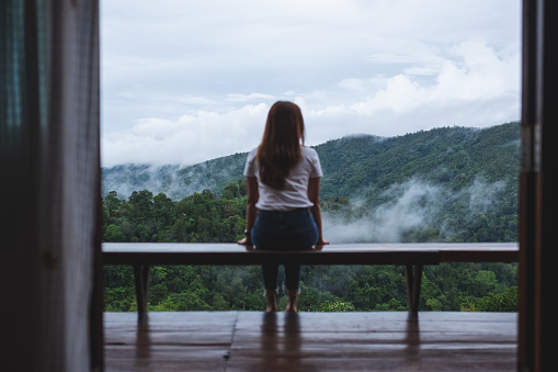 istock Rear view image of a woman sitting on wooden balcony while watching a beautiful mountains and nature view on foggy day 1402998911