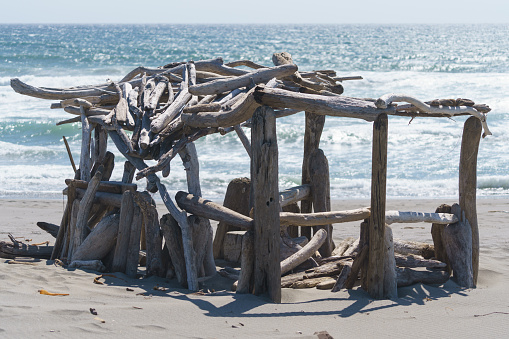 Driftwood hut, fort or house on beach on sunny day by ocean.