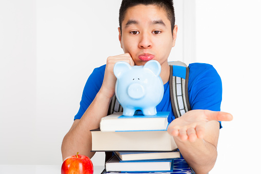Pouty asian University student in blue shirt with hand palm open asking for college donation sitting at desk