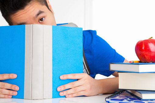 Vietnamese adult student in blue shirt hiding behind book on white table at home