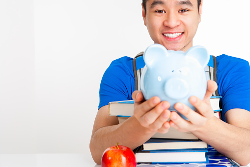 Smiling Vietnamese University student in blue shirt holding blue piggy bank sitting at table