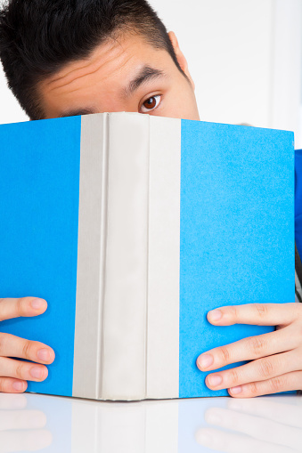 Vietnamese adult student peeking over blue textbook sitting at desk