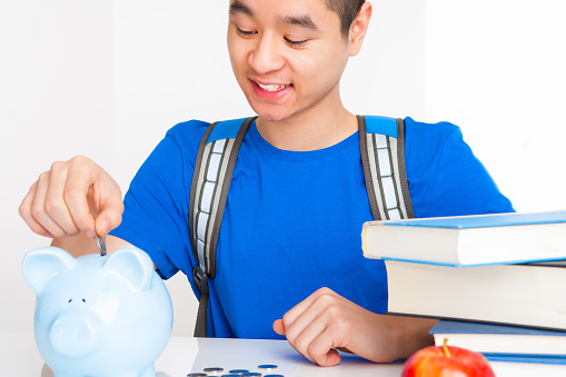 Smiling asian University student in blue shirt inserting money into piggy bank sitting at desk.