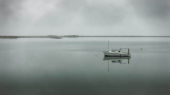 A monochrome shot of a boat at Motueka seafront on a grey gloomy evening, a moored motor cruiser, the Motueka sandspit in the background, Tasman region, Aotearoa / New Zealand.