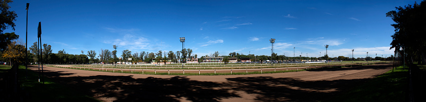 Independence Hippodrome, panoramic view, made with the union in computer of seven individual photos. The photos were taken from outside to the Racetrack, just next to the property, from the public park that surroud the place. The image shows the track at foregound and the tribunes at the back, on a sunny day.\nLocation: Rosario city, SantaFe province, Argentina. Photo date: May 8, 2022