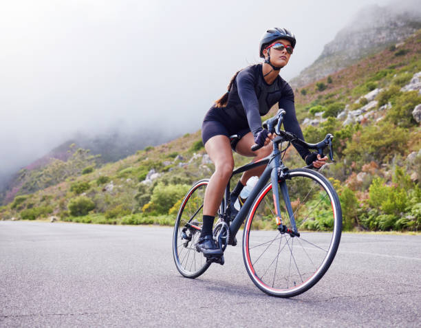 外を自転車で走る一人の運動選手の若い女性。ヘルメットとメガネをかけたスポーティなフィット感の女性は、運動のために山沿いの道路で自転車に乗っています。ワークアウトとトレーニ� - cycling ストックフォトと画像