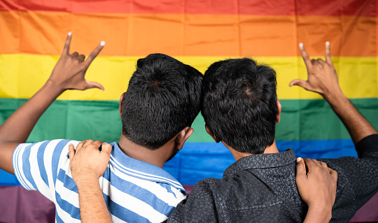 back view of young male couple showing love sign in front of pride or rainbow flag, lgbtq concept.