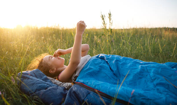 the girl is dissatisfied with scratching mosquito bites, child sleeps in a sleeping bag on the grass in a camping trip. eco-friendly outdoor recreation, summer time. sleep disturbance, repellent. - mosquito child bug bite scratching imagens e fotografias de stock