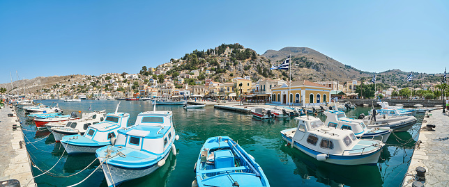 Symi, Greece - July 05 2017: Moored tourist and fishing boats at pier in port of island Symi in Greece. Vessels drift on turquoise water against small multicolored houses on hills