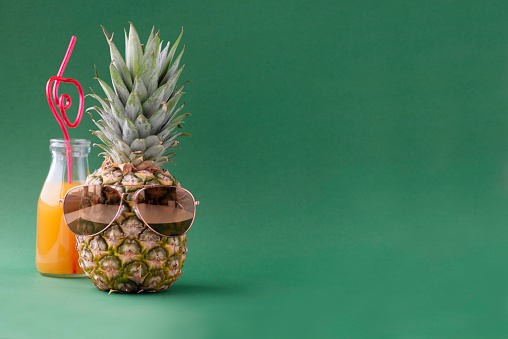A bottle of fruit juice and a pineapple wearing sunglasses in front of green background.