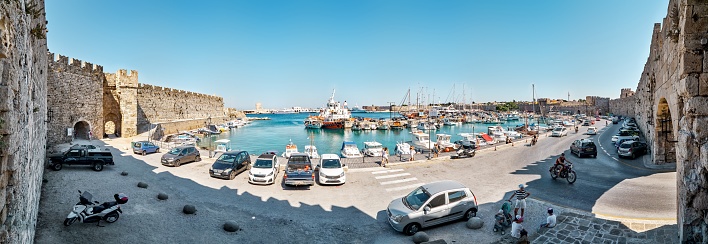 Rhodes, Greece - July 08 2017: Ancient Rhodes city port and fortress walls in Greece. Boats and yachts moored to pier along bay seafront with parked cars near ancient castle