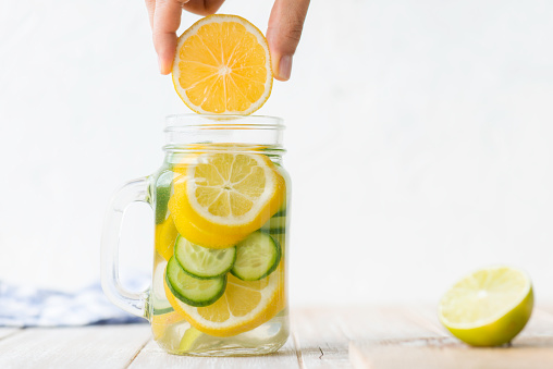 Infused water bottle with an abundance of lemon and cucumber slices on white wooden table. Hand of an unrecognizable person is adding lemon slice