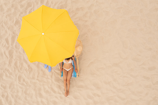 Woman resting in sunbed under yellow beach umbrella at sandy coast, aerial view. Space for text