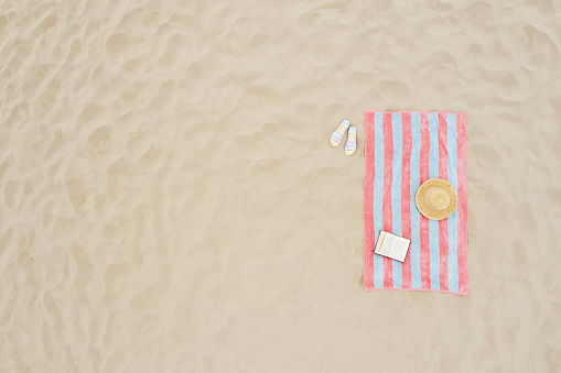 Striped beach towel, book, straw hat and flip flops on sand, aerial view. Space for text