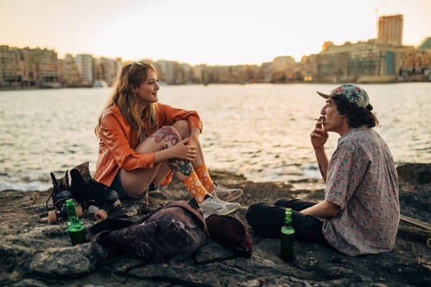 A multiracial man and a woman enjoy relaxing on the rocky shore. They drink beer and smoke weed Two cheerful best friends smoke weed and drink a refreshing beer while sitting on a rocky shore near the sea golden hour drink stock pictures, royalty-free photos & images