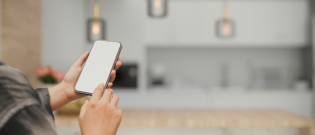 A mockup screen of a smart mobile phone or cellphone white blank screen in a woman's hand over blurred modern luxury white kitchen room in the background. 3d rendering, 3d illustration