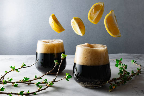 Two low full glasses with a dark carbonated drink with a high foam, beer or kvass, flying lemon slices and a young currant sprig stock photo