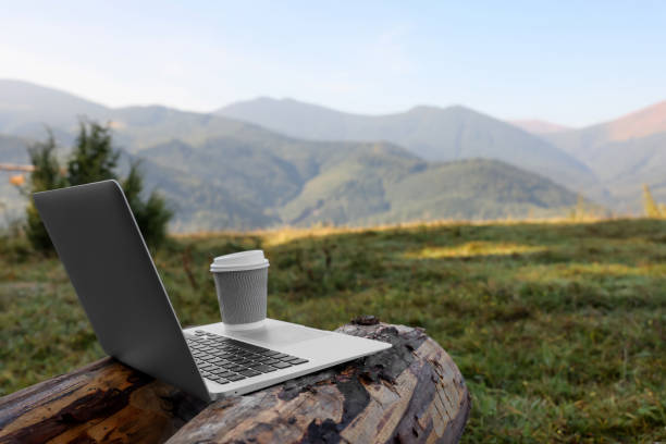 istock Modern laptop and coffee cup on log in mountains, space for text. Working outdoors 1403030089